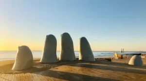 Sculpture d'une main sortant du sable sur la plage devant l'océan | itinerares